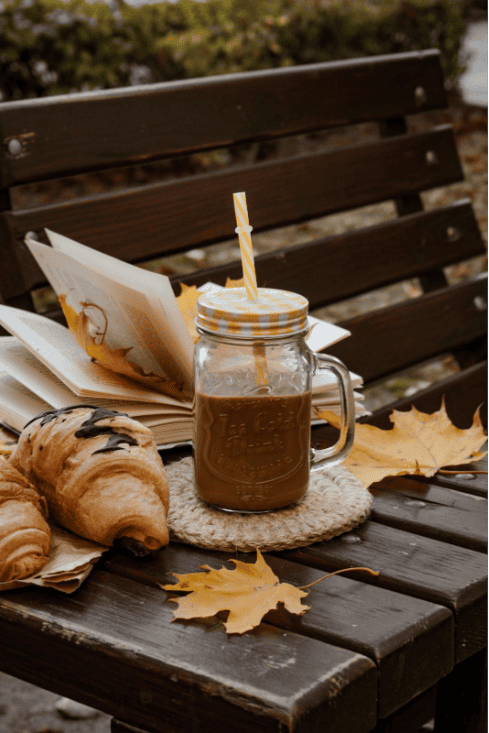 Cup of iced coffee in a mason jar on a wooden bench with a book, croissant, and autumn leaves.